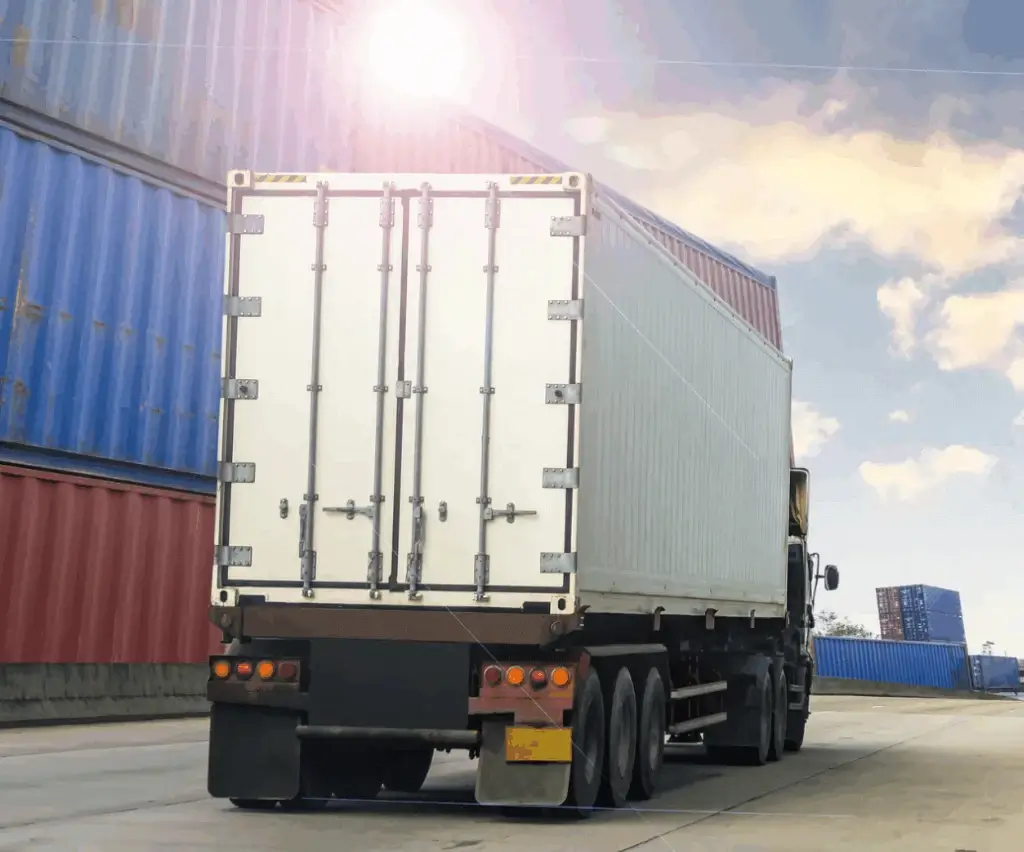 Why Importers Should Book Their Own Freight 9 Container freight truck pulling out of a shipping yard surrounded by stacked red and blue cargo containers under a bright sky.