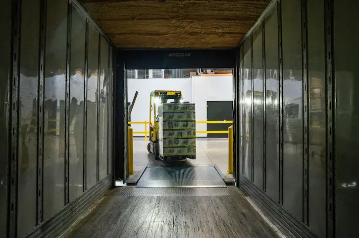 Warehouse dock view of a forklift moving a wrapped pallet into a trailer for FDA import requirements clearance.
