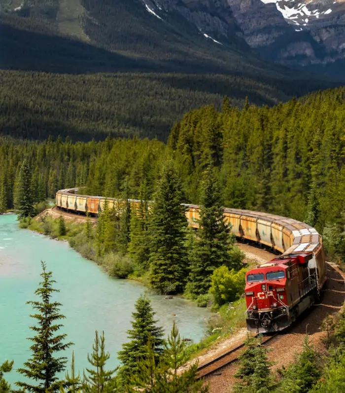 Red diesel locomotive pulling a long freight train along a winding track beside a turquoise river through a forested valley.