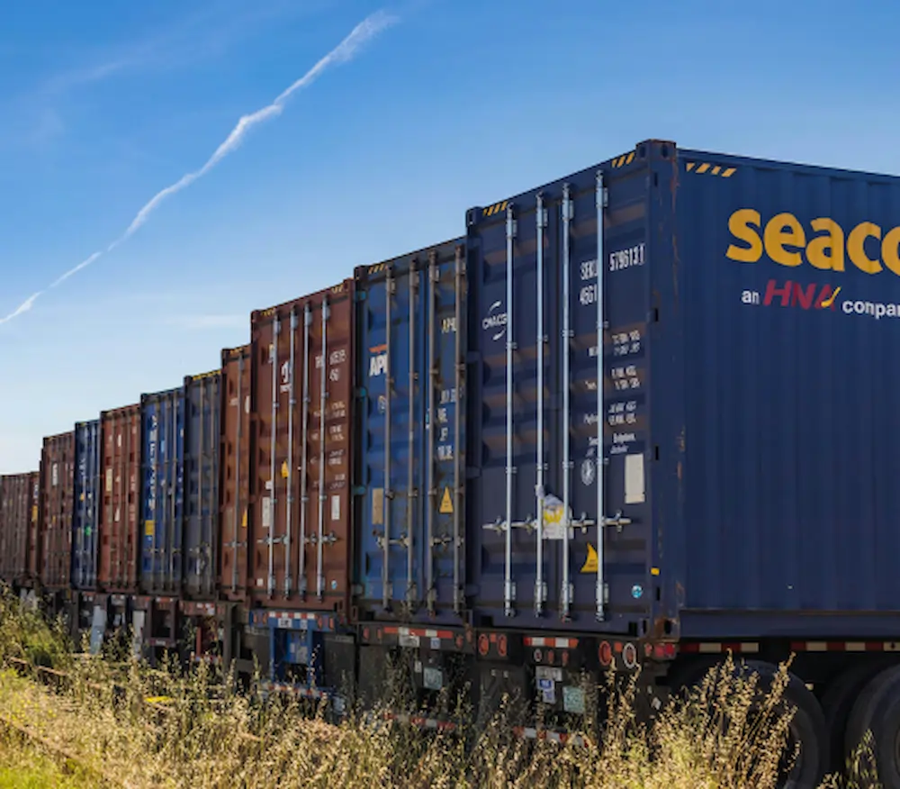 Line of colorful shipping containers on flatbed train cars beside a grassy field under a clear blue sky, with Seaco branding visible on the right container.