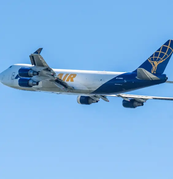 Atlas Air cargo jet in flight, white fuselage with orange 'AIR' branding and blue tail with globe logo when viewed against clear blue sky.
