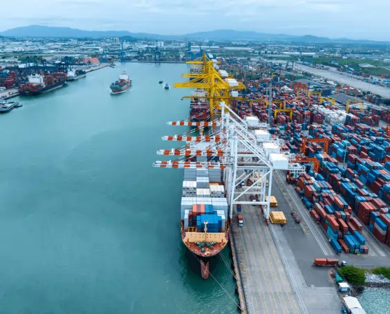 Aerial view of a busy shipping port with stacked containers and large yellow port cranes unloading a cargo ship at the dock.