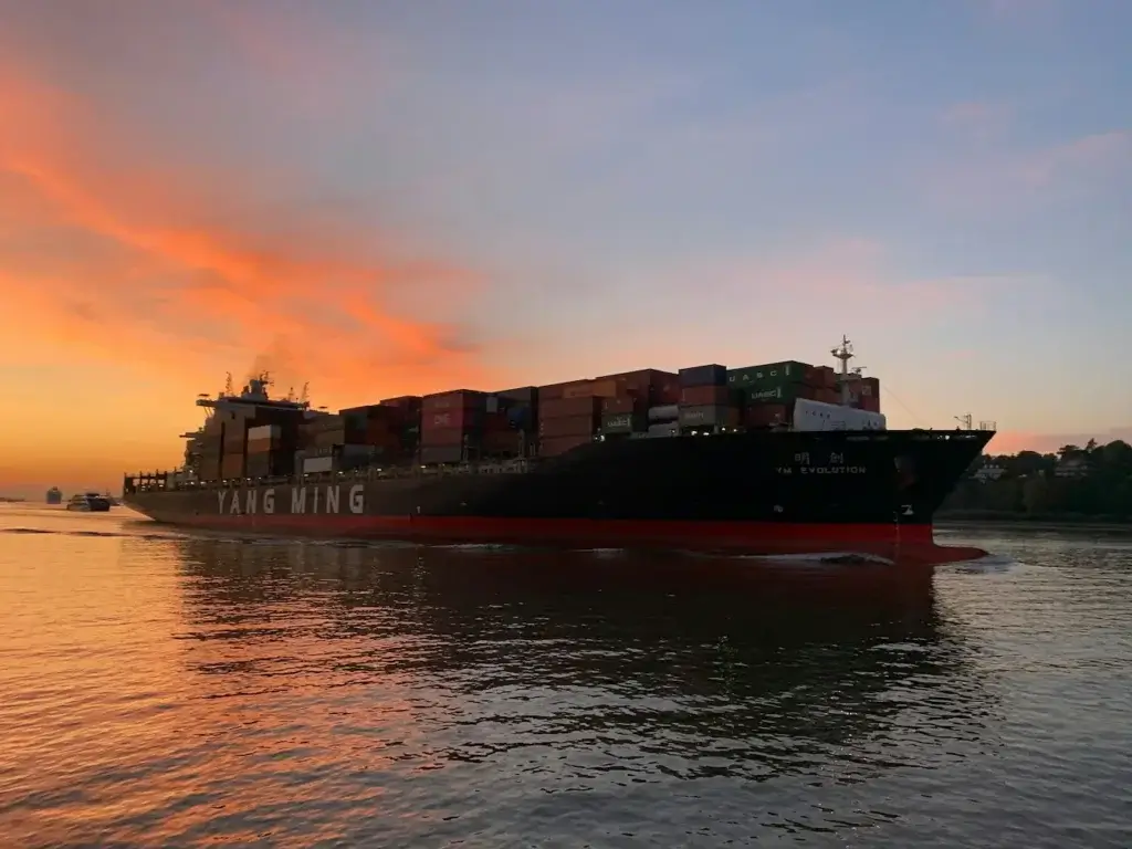 A Guide to Shipping Oversized and Heavy Cargo Internationally 2 A cargo ship loaded with oversized shipping containers sailing at sunset, with a vibrant orange sky reflected on the water.