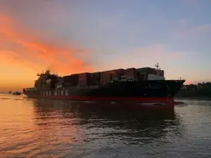 A cargo ship loaded with oversized shipping containers sailing at sunset, with a vibrant orange sky reflected on the water.