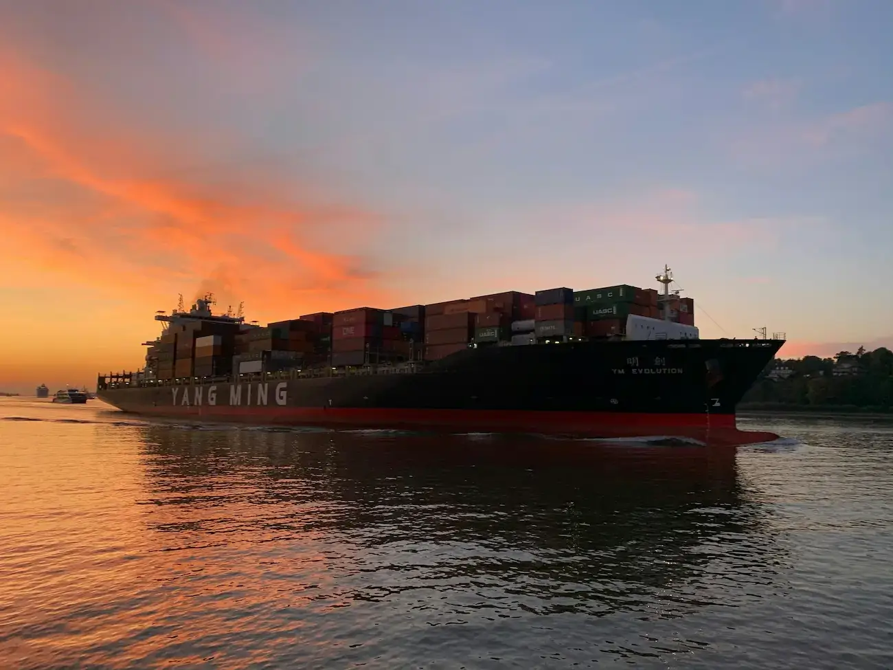 A cargo ship loaded with oversized shipping containers sailing at sunset, with a vibrant orange sky reflected on the water.