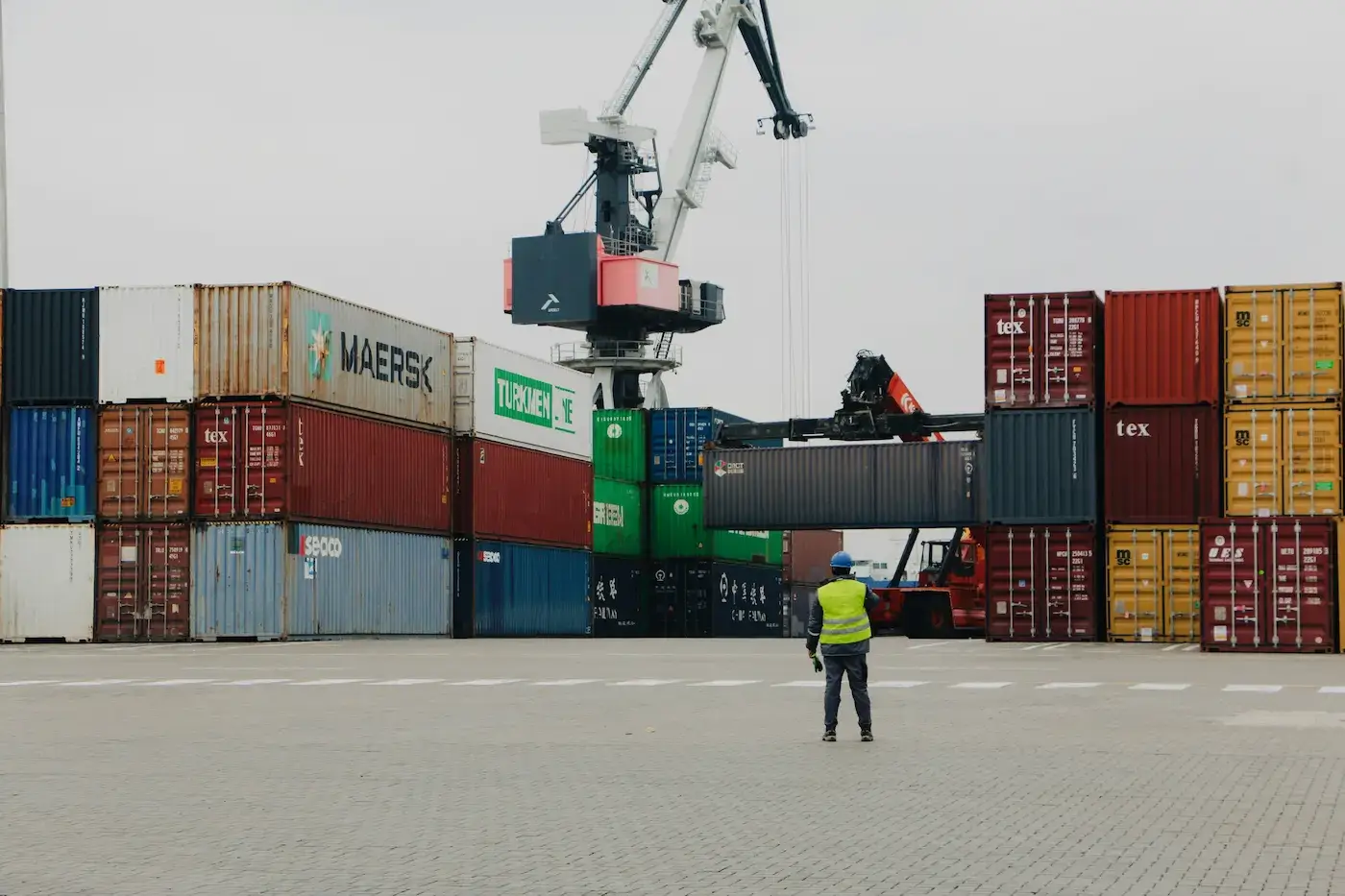 Custom clearance port worker in a yellow safety vest and hard hat overseeing a crane lifting shipping containers at a busy freight terminal stacked with Maersk and Turkmen Line cargo containers.
