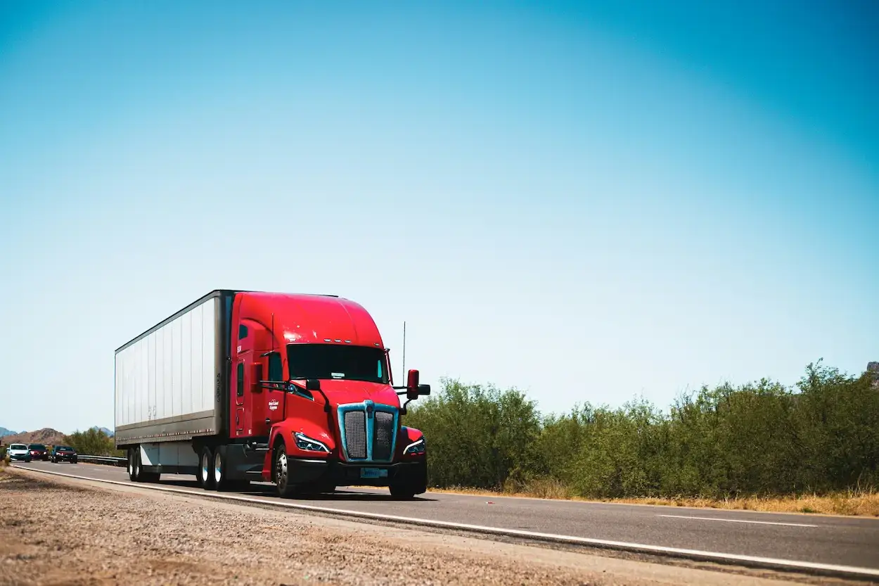 Red dry van full sized truck driving on a desert highway under a clear blue sky.