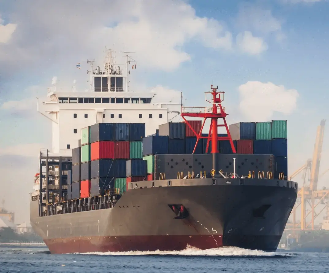 Front view of a container ship loaded with colorful cargo containers navigating open water toward a port, representing Jade International's ocean export and international freight forwarding services.