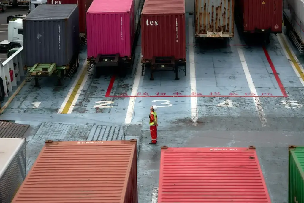 Importer of record dock worker in safety gear inspecting numbered loading lanes filled with colorful shipping containers on a cargo vessel deck.