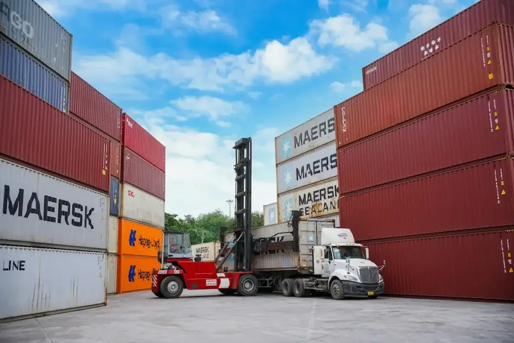 Incoterms shipping yard with a forklift loading a Maersk Sealand container onto a truck, surrounded by stacked Maersk and Hapag-Lloyd cargo containers under a blue sky.