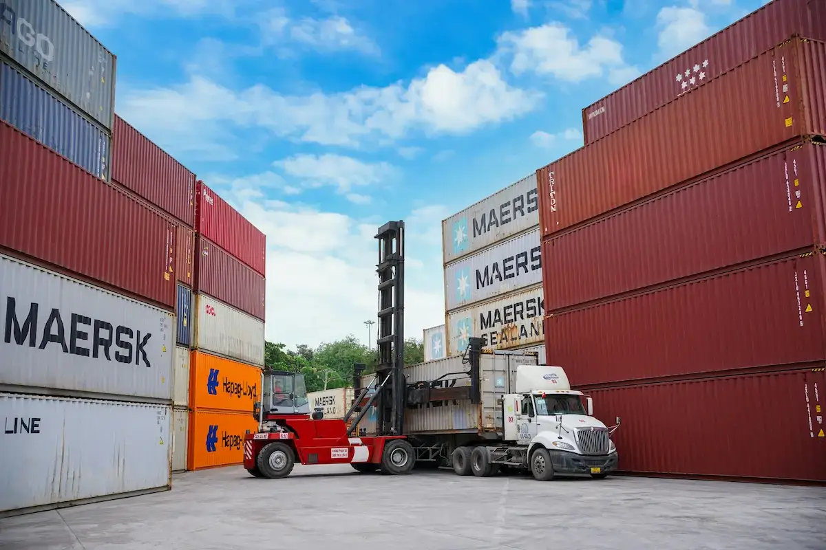 Incoterms shipping yard with a forklift loading a Maersk Sealand container onto a truck, surrounded by stacked Maersk and Hapag-Lloyd cargo containers under a blue sky.
