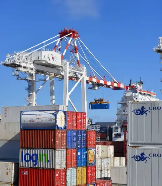 Port crane loading a cargo container onto a ship, with medical shipment containers stacked at the freight terminal
