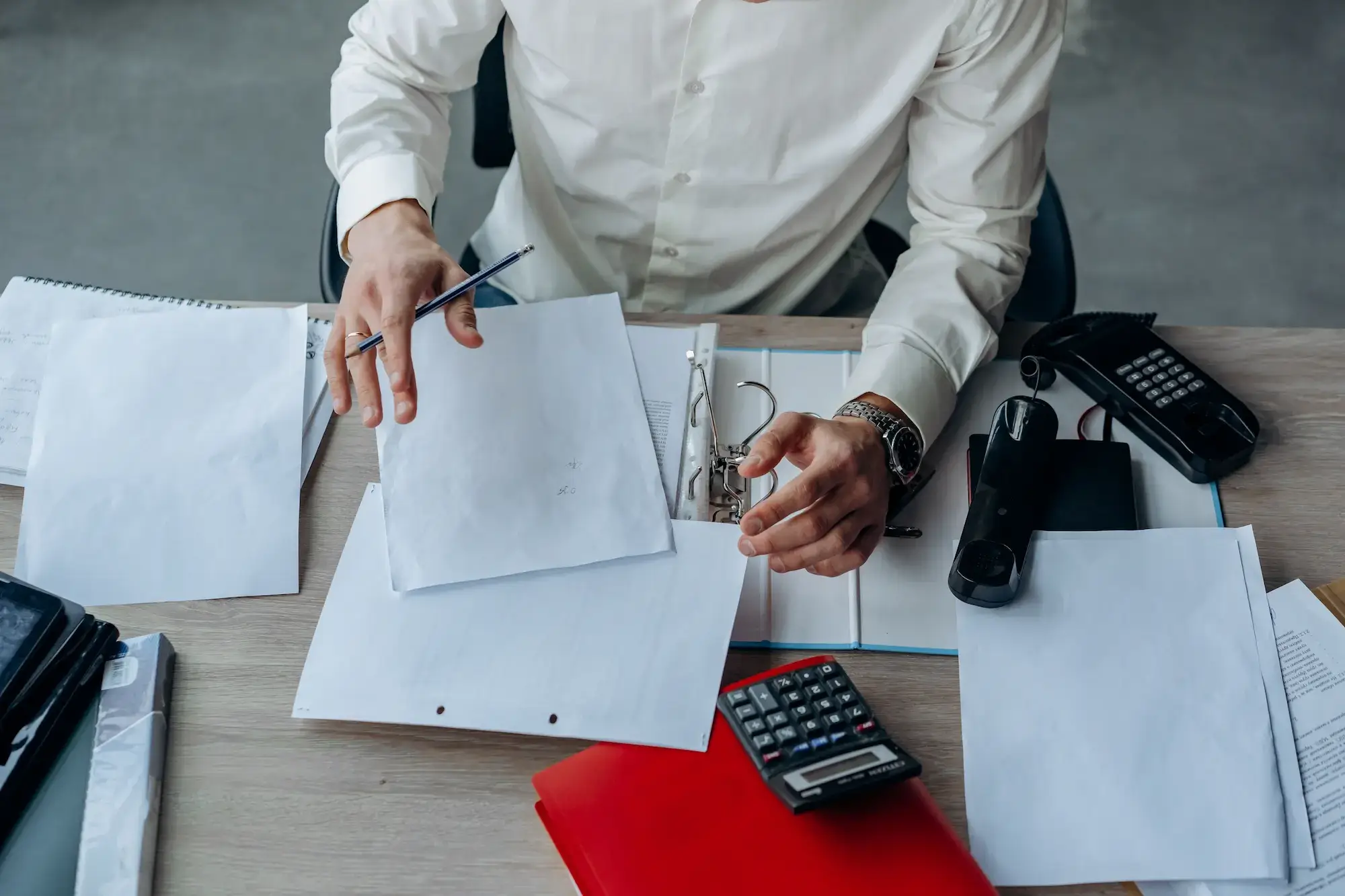 Overhead view of a person at a desk sorting through paperwork, binders, and a calculator, illustrating documentation for international shipping.