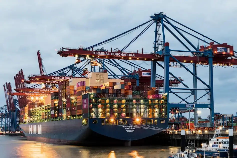 HMM Nuri container ship loaded with cargo at Terminal Burchardkai under gantry cranes at dusk, illustrating freight forwarder operations.