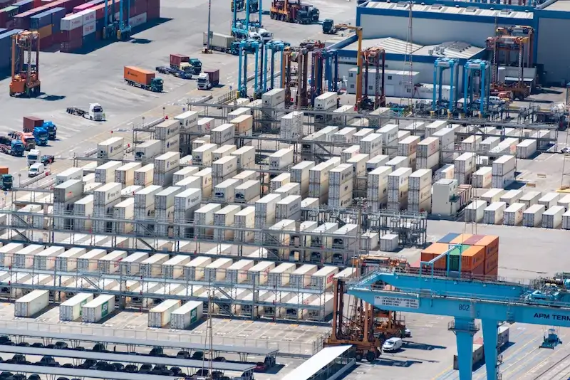 Aerial view of a container freight station with stacked shipping containers, gantry cranes, and trucks moving cargo across the terminal yard.